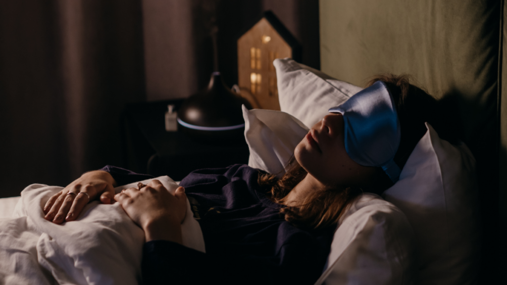 A person sleeping in bed, lying on their back under the covers in a dimly lit room, wearing a blue silk sleep mask.