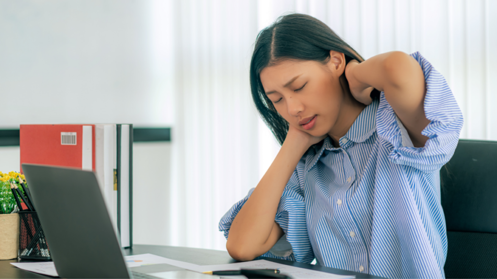 A person wearing a blue button up shirt, sitting at a desk at work in front of a laptop, eyes closed and both arms raised, holding the back of their neck. Their face shows signs of discomfort.