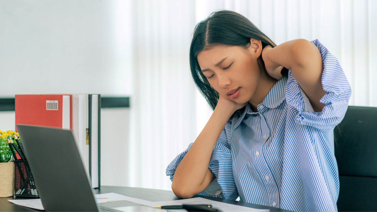 Woman experiencing tech neck sits at an office desk in front of a computer in pain.