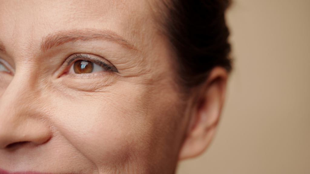 A close-up of a middle-aged woman's face, focusing on her brown eye and the surrounding skin. She has a warm expression with slight wrinkles around her eye. The background is blurred and neutral-toned.