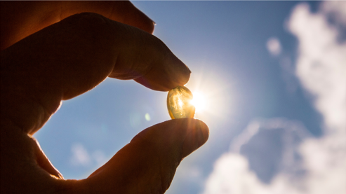 A hand holds a translucent vitamin D capsule up to the sunlight, with the sun shining through the soft gel against a backdrop of a bright blue sky and scattered clouds.