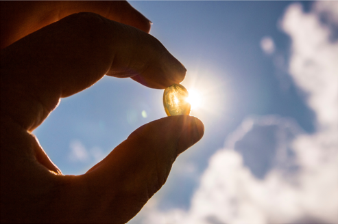 A hand holds a translucent vitamin D capsule up to the sunlight, with the sun shining through the soft gel against a backdrop of a bright blue sky and scattered clouds.