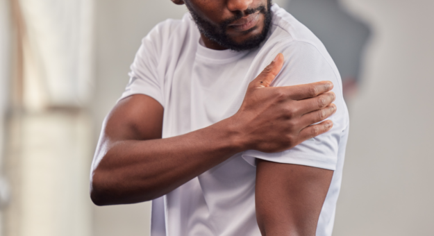 A man wearing a white athletic shirt holds his shoulder in discomfort, possibly experiencing shoulder pain. His facial expression suggests concern as he looks at the affected area. The background is blurred, indicating an indoor setting.